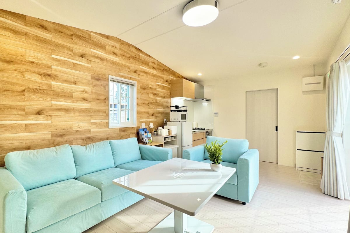 A modern living room with a wooden accent wall, a light blue sofa, a white coffee table, and a kitchen area with a microwave and cabinets.