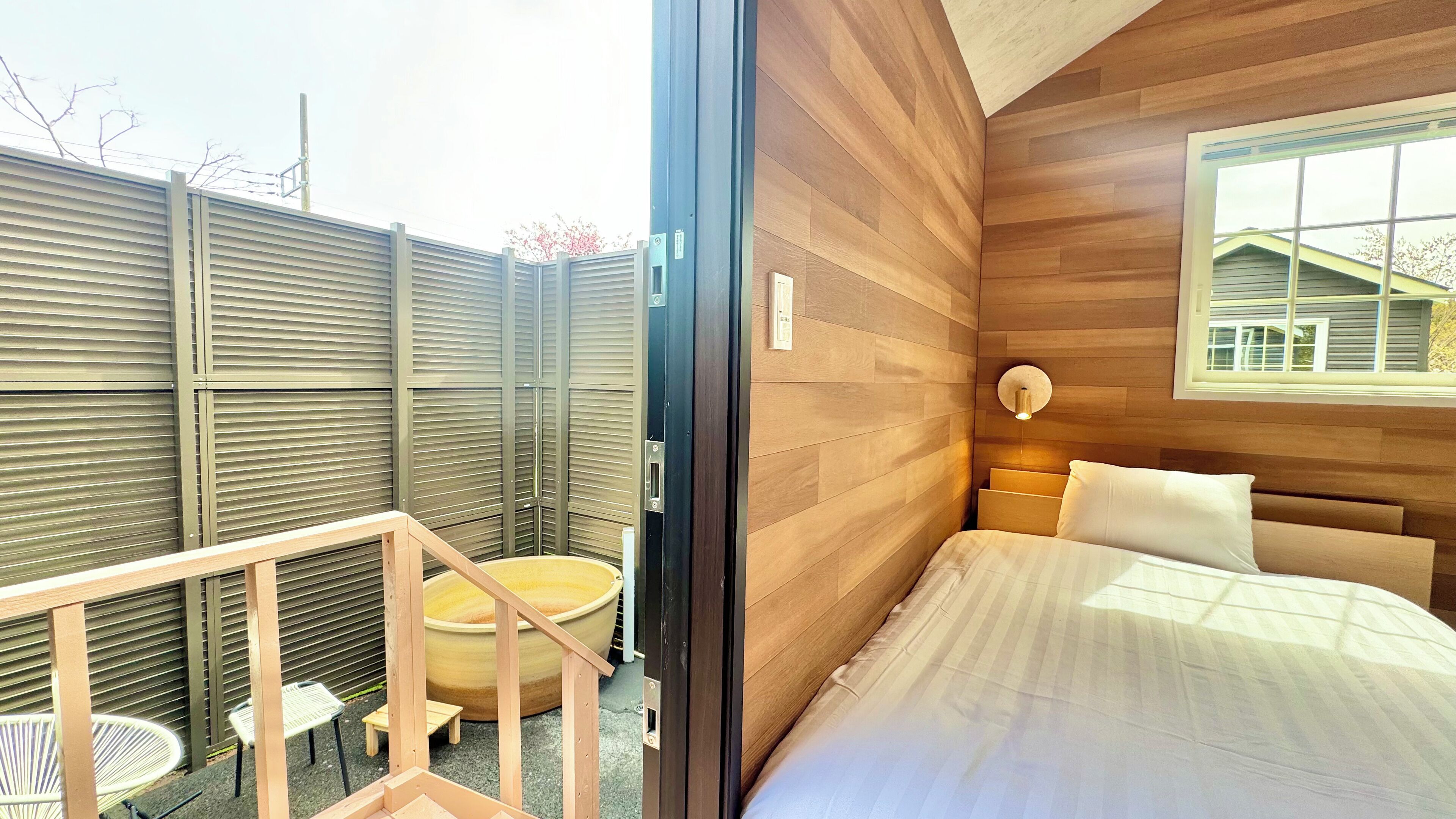 A bedroom with a bed, a window, and a view of a house. There is a small table and chairs outside the room.