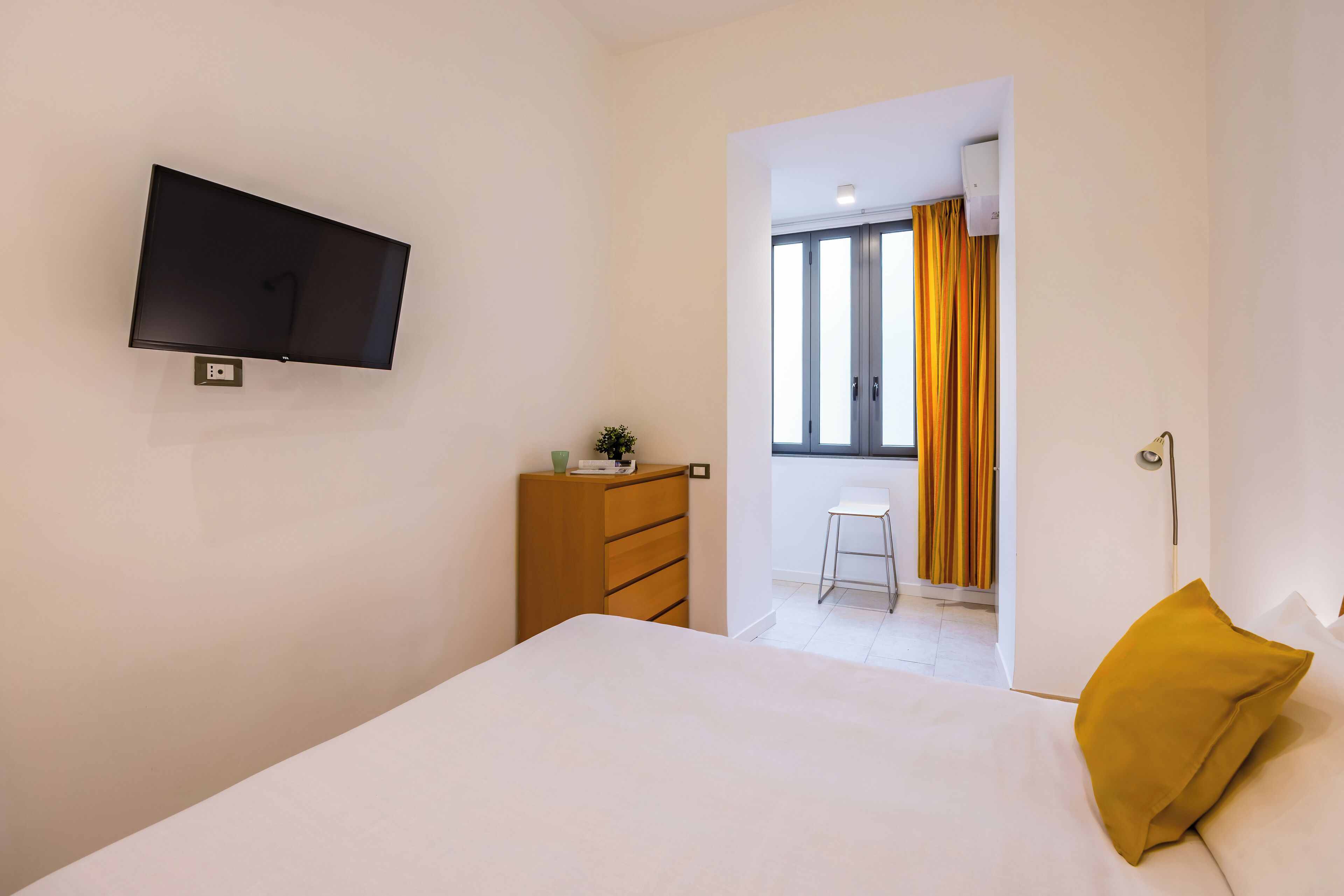 Een slaapkamer met een bed, een aan de muur bevestigde televisie, een houten dressoir en een raam met gordijnen.
