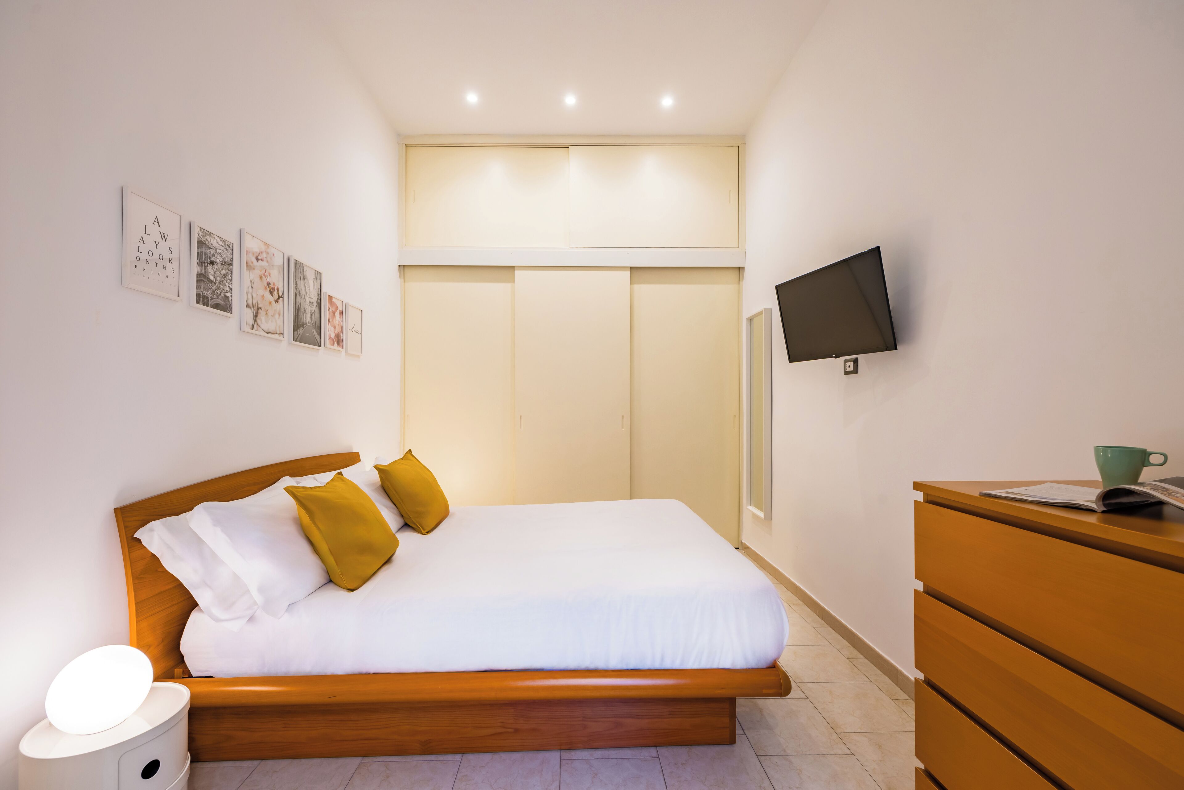 Une chambre avec un lit en bois, une literie blanche, une table de chevet avec une lampe, une commode et un téléviseur à écran plat fixé au mur.