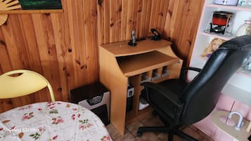 A compact room with a desk, a chair, a small table, and a shelf with various items.