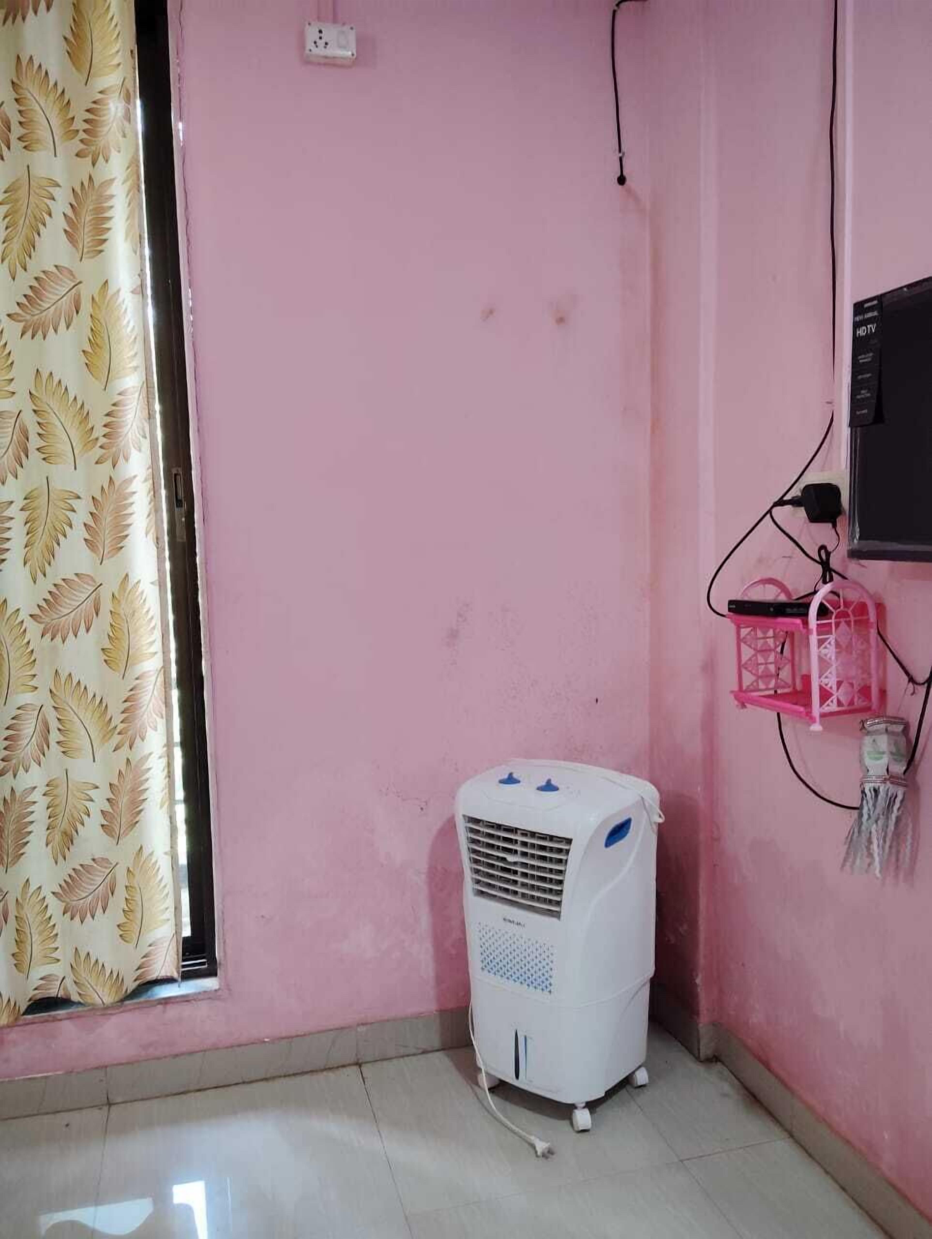 Een kamer met roze wanden, een raam met een gordijn met bladmotief, een witte mobiele airconditioner en een roze speaker aan de muur.