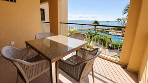 Un balcón con una mesa de vidrio y sillas de mimbre, con vista a una piscina y palmeras.