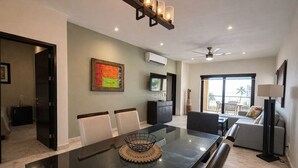 A modern living room with a glass dining table, a sofa, and a TV. There is a painting on the wall and a ceiling fan. - Fully remodeled two-bedroom ocean view condo at ELFARO by DPM (Playa del Carmen)