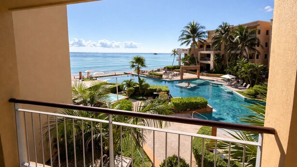 Vista de una piscina con un océano azul cristalino al fondo, rodeada de palmeras y un edificio con balcones.