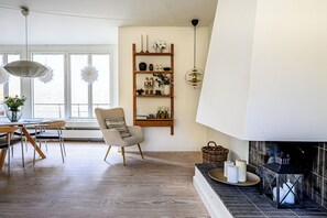 A modern living room with a wooden floor, a dining table with chairs, a sofa, a fireplace, and a wall-mounted shelf with decorative items.