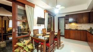 A dining area with a wooden table and chairs, a television mounted on the wall, and a kitchen with wooden cabinets. - 9BR Private Pool Sanur Beach Villa (3units villa) (Bali)