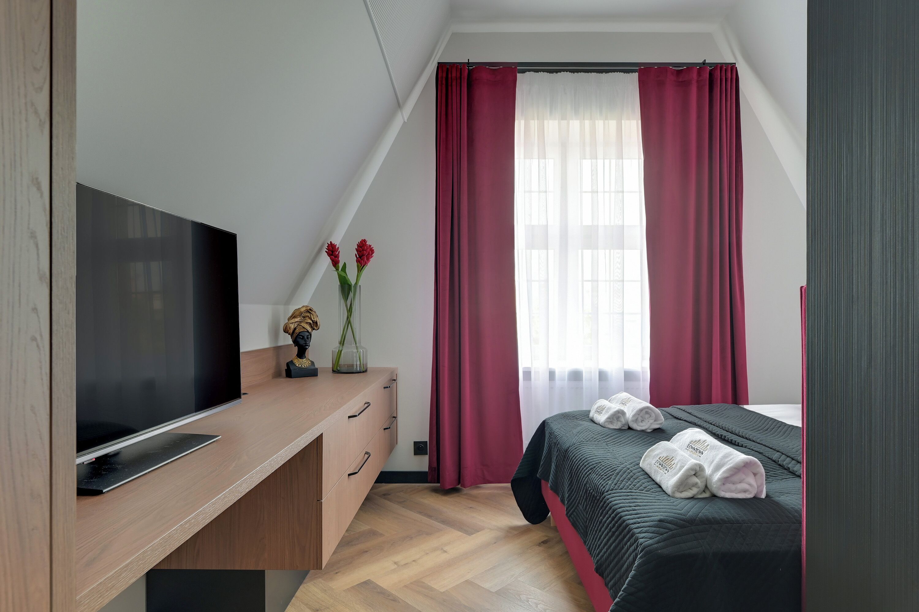 A modern bedroom with a flat-screen TV, wooden dresser, and a bed with neatly folded towels.