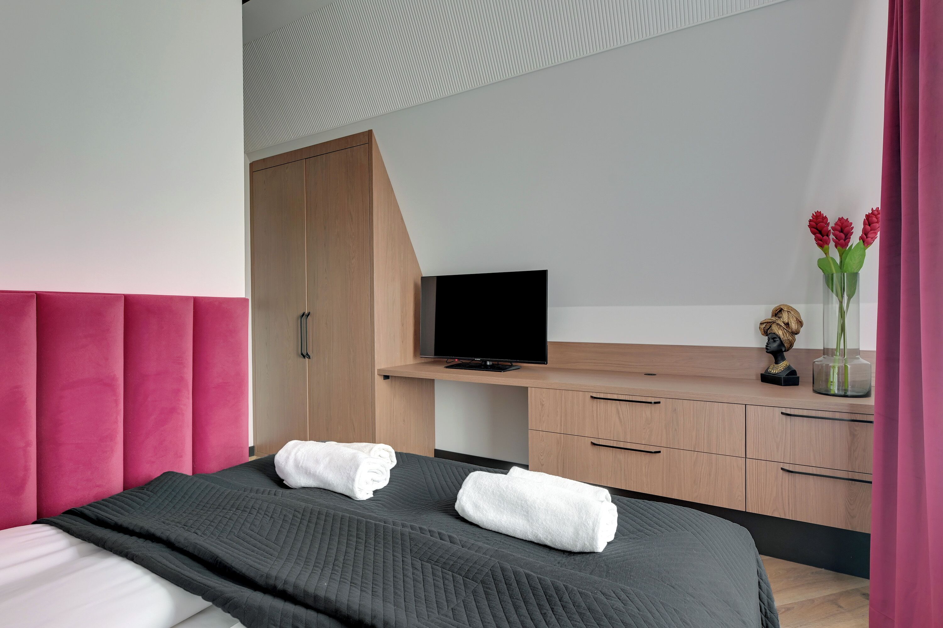 A modern hotel room with a bed, a desk with a flat-screen TV, wooden cabinets, and a vase with red flowers.