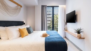 Una habitación de hotel moderna con una cama grande, un televisor montado, una ventana con vistas a edificios y un estante pequeño con una planta.