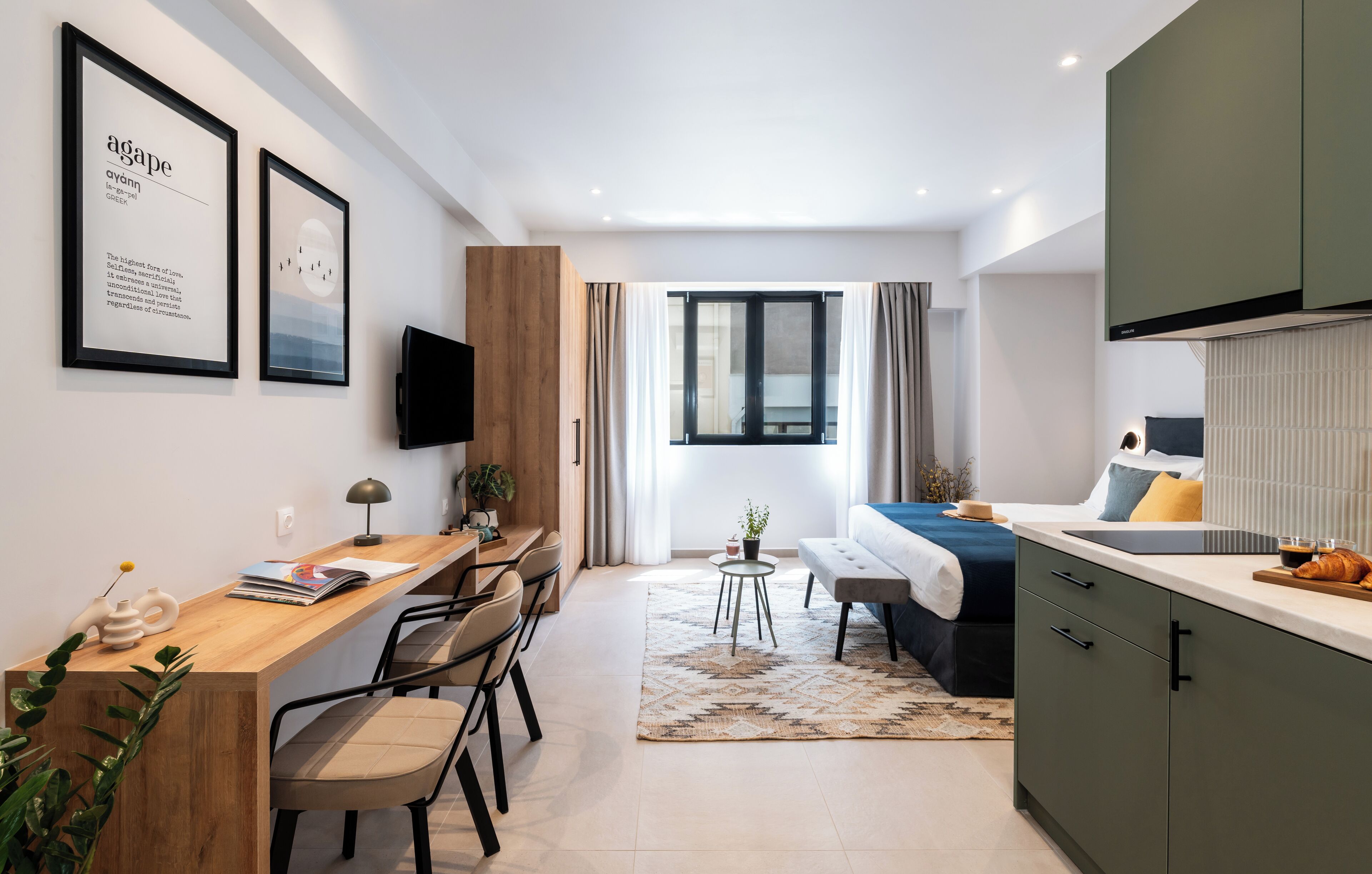 A modern, well-lit room with a wooden desk, a bed with a blue bedspread, a dining area, and a kitchen with a built-in oven.