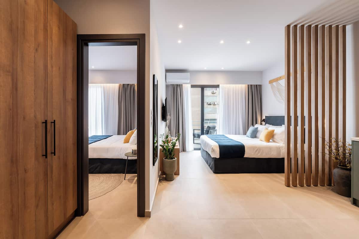 A modern hotel room with two beds, a wooden wardrobe, a large potted plant, and a view of the city.