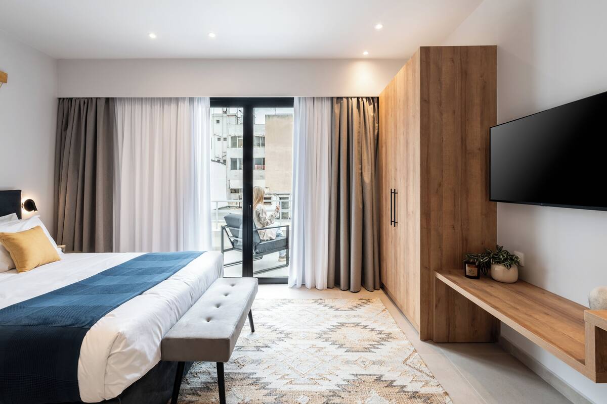 A modern hotel room with a large bed, a bench, a TV, a wooden wardrobe, and a balcony with curtains.