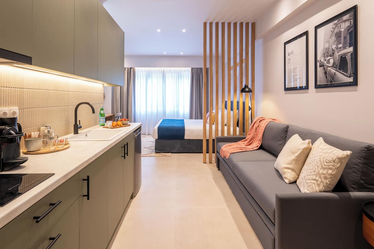 A modern hotel room with a kitchenette, a bed with a blue headboard, a grey sofa with patterned cushions, and two framed pictures on the wall.