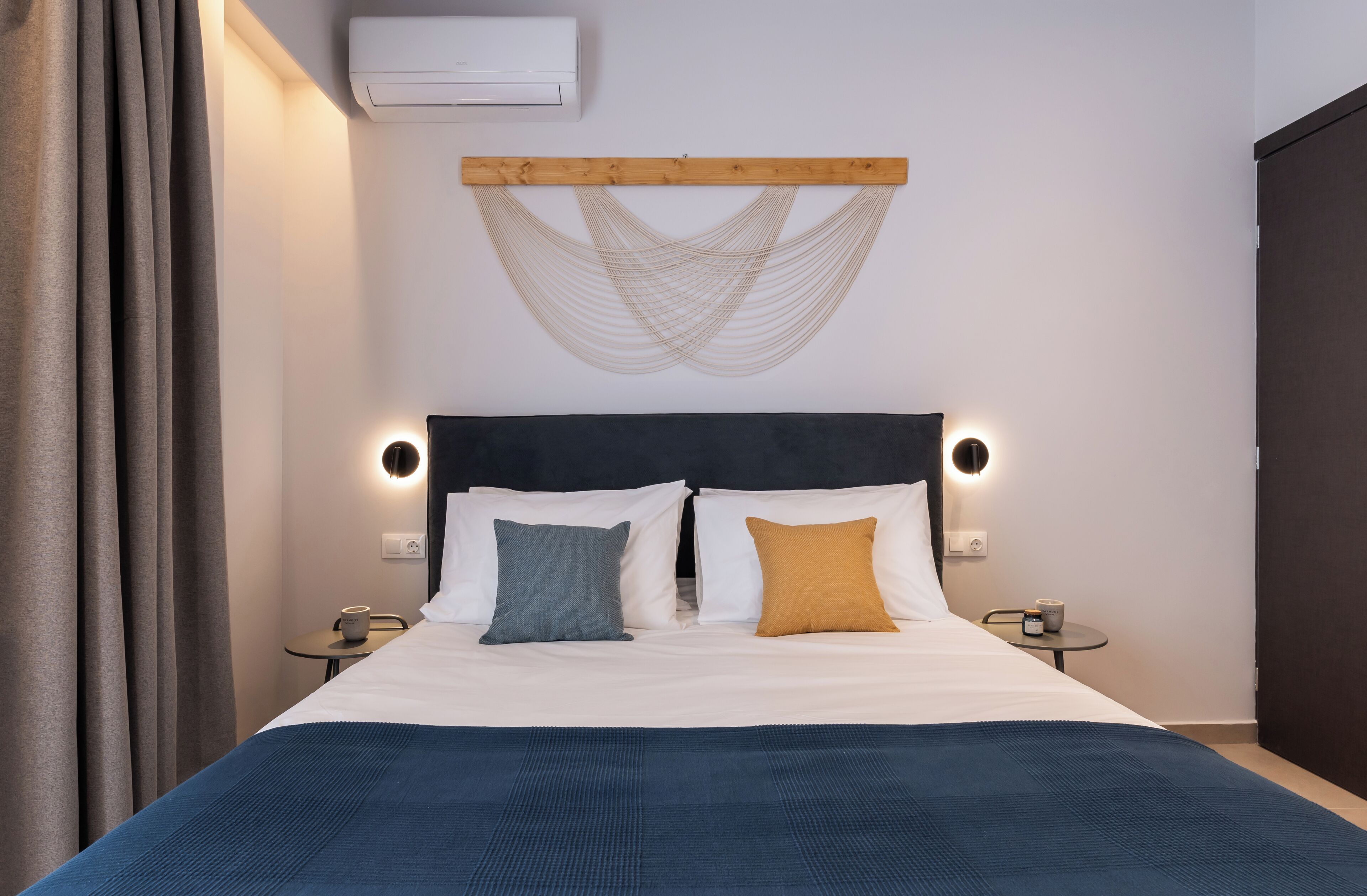 A neatly made bed with a blue blanket, two pillows, and a wooden headboard. There are bedside tables with lamps and a wall-mounted air conditioner.