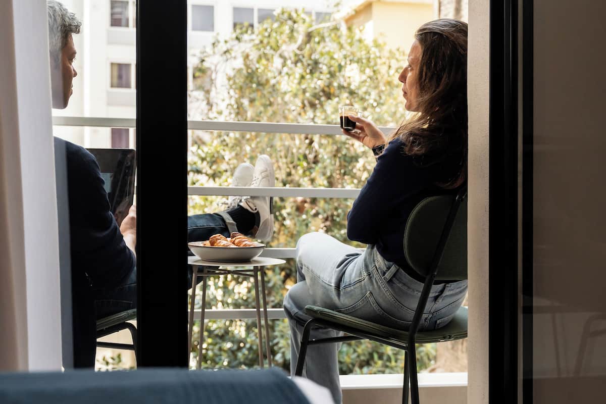 A balcony with two people seated, one holding a glass. A table with a bowl of food is visible.