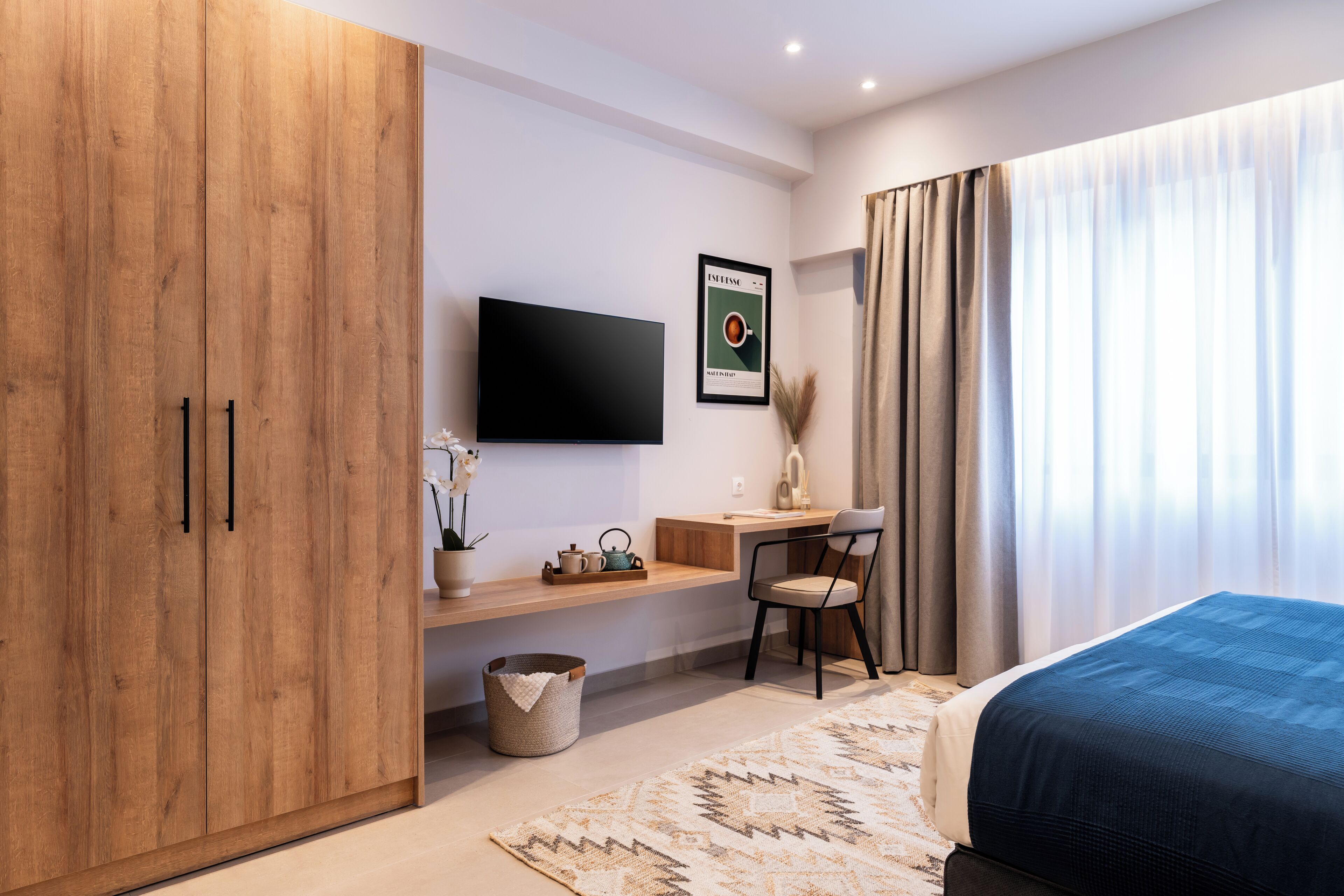 A modern bedroom with a large wardrobe, a flat-screen TV, a desk with a chair, and a bed with a blue cover.