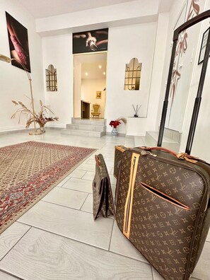 Ein geräumiges Zimmer mit einem großen Spiegel vom Boden bis zur Decke, einer Louis Vuitton Tasche, einem roten Teppich mit Muster und einer Vase mit roten Blumen.