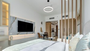 A modern hotel room with a large bed, a flat-screen TV mounted on the wall, a small dining area, and a visible kitchenette.