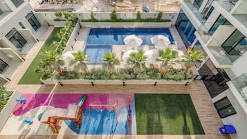 A rooftop pool area with a blue pool, palm trees, and a children's play area with a slide and climbing structure.