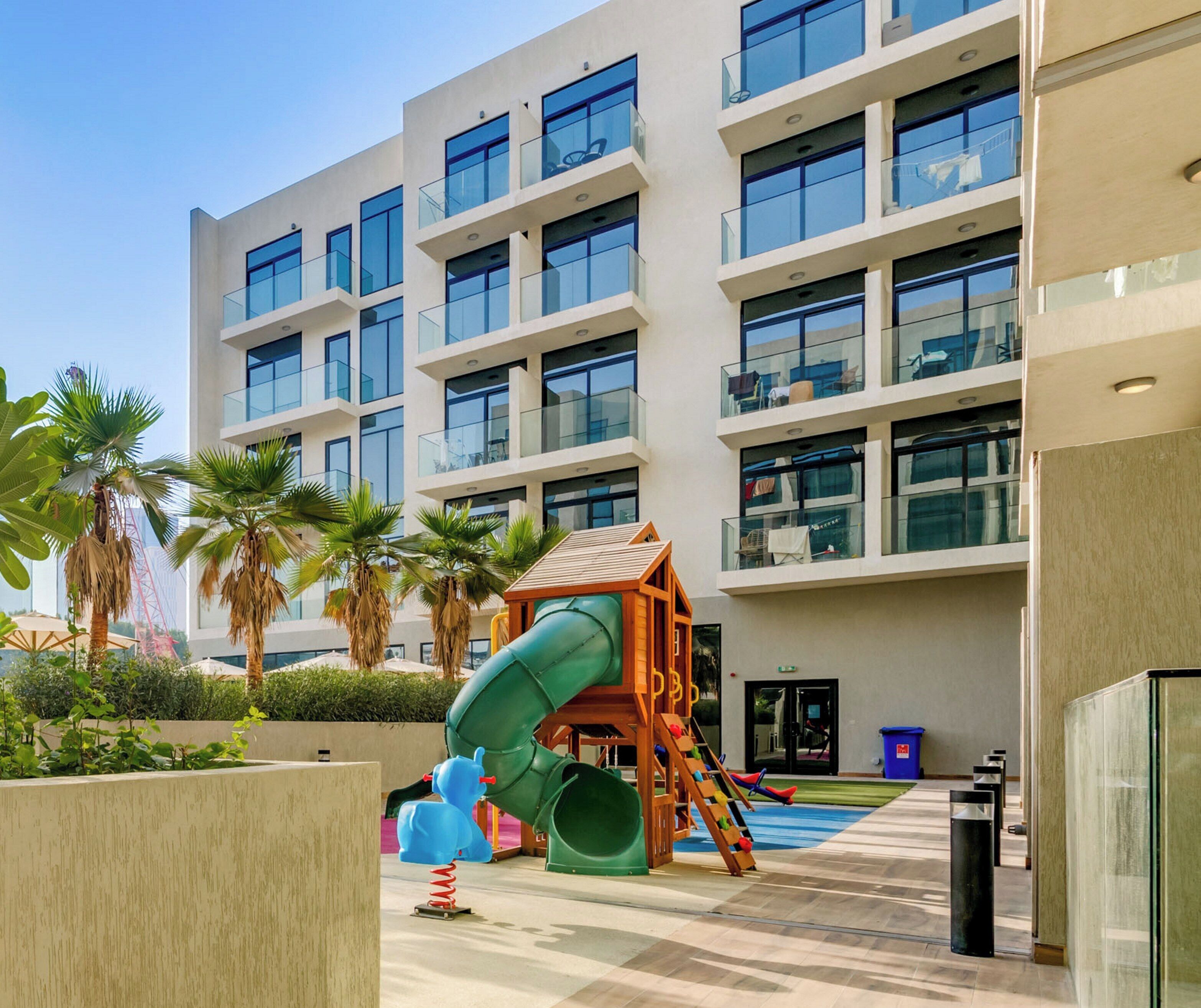 Un bâtiment moderne doté de nombreux balcons, d’un espace de jeux pour enfants avec un toboggan et de palmiers.