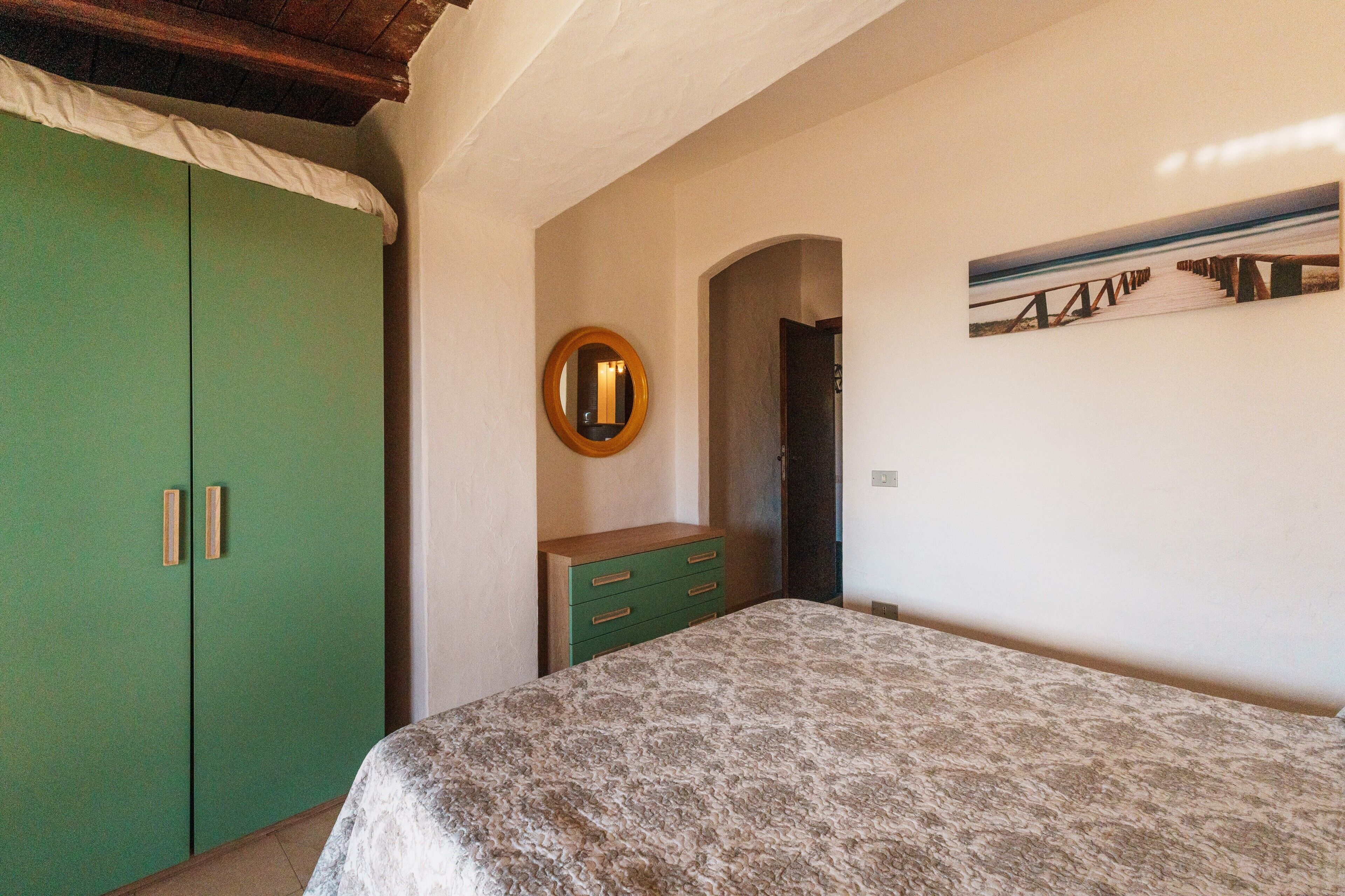 A bedroom with a bed, a green wardrobe, a dresser, a mirror, and a painting on the wall.
