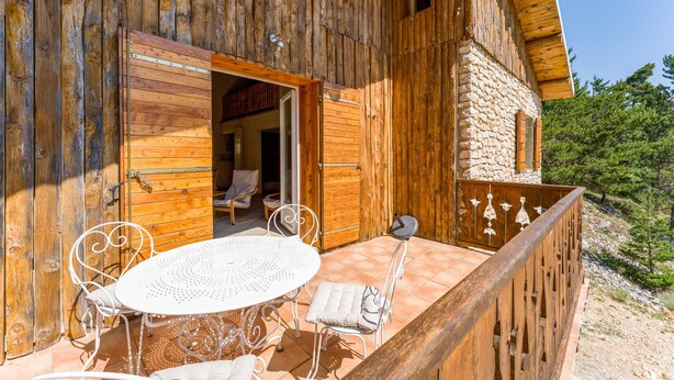 Eine Holzhütte mit Steinwand, einem Balkon mit Metallgeländern und einem runden Tisch mit Stühlen.