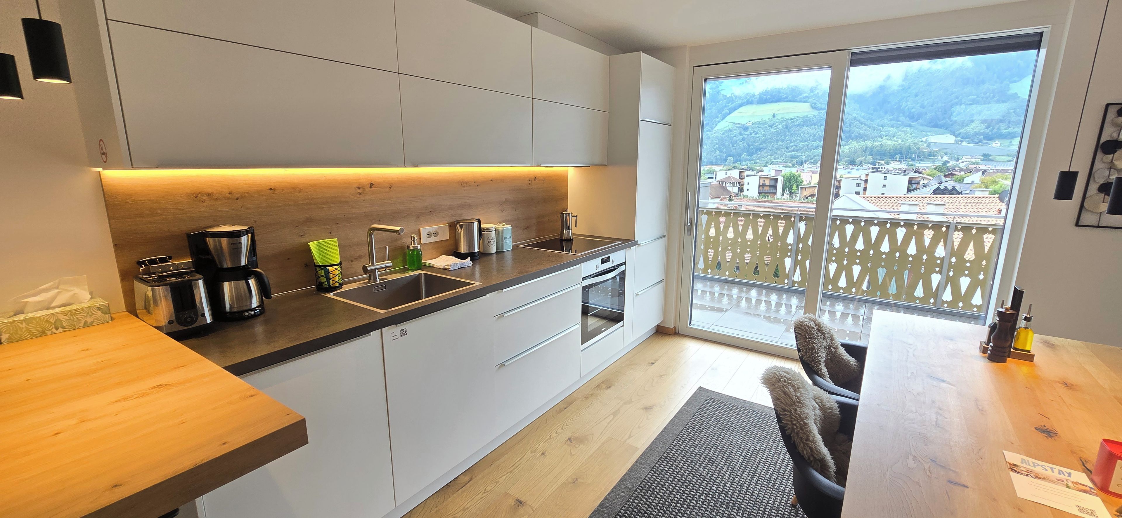 Una cucina moderna con un'ampia finestra, un'area pranzo con tavolo in legno e vista su un paesaggio montuoso.