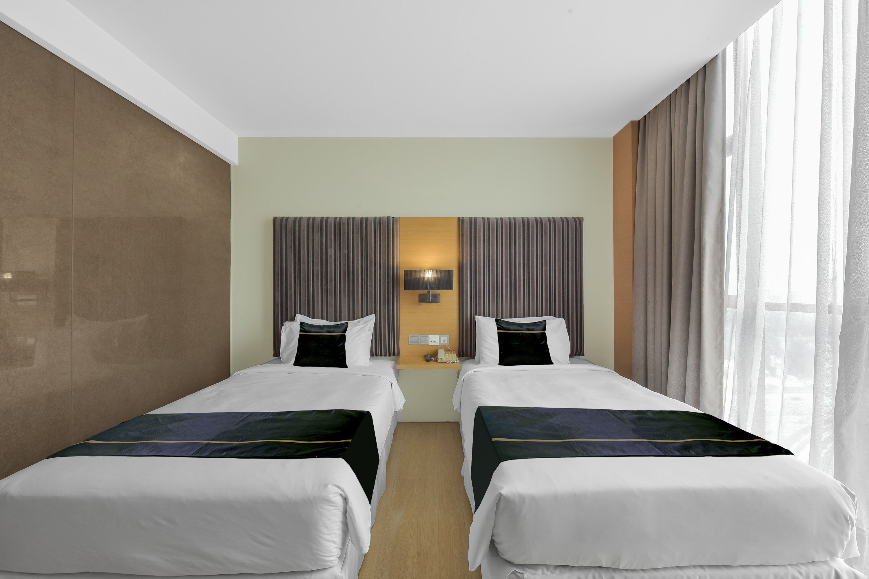 Quarto de hotel com duas camas, uma mesa de cabeceira, uma luminária e uma janela grande com cortinas.