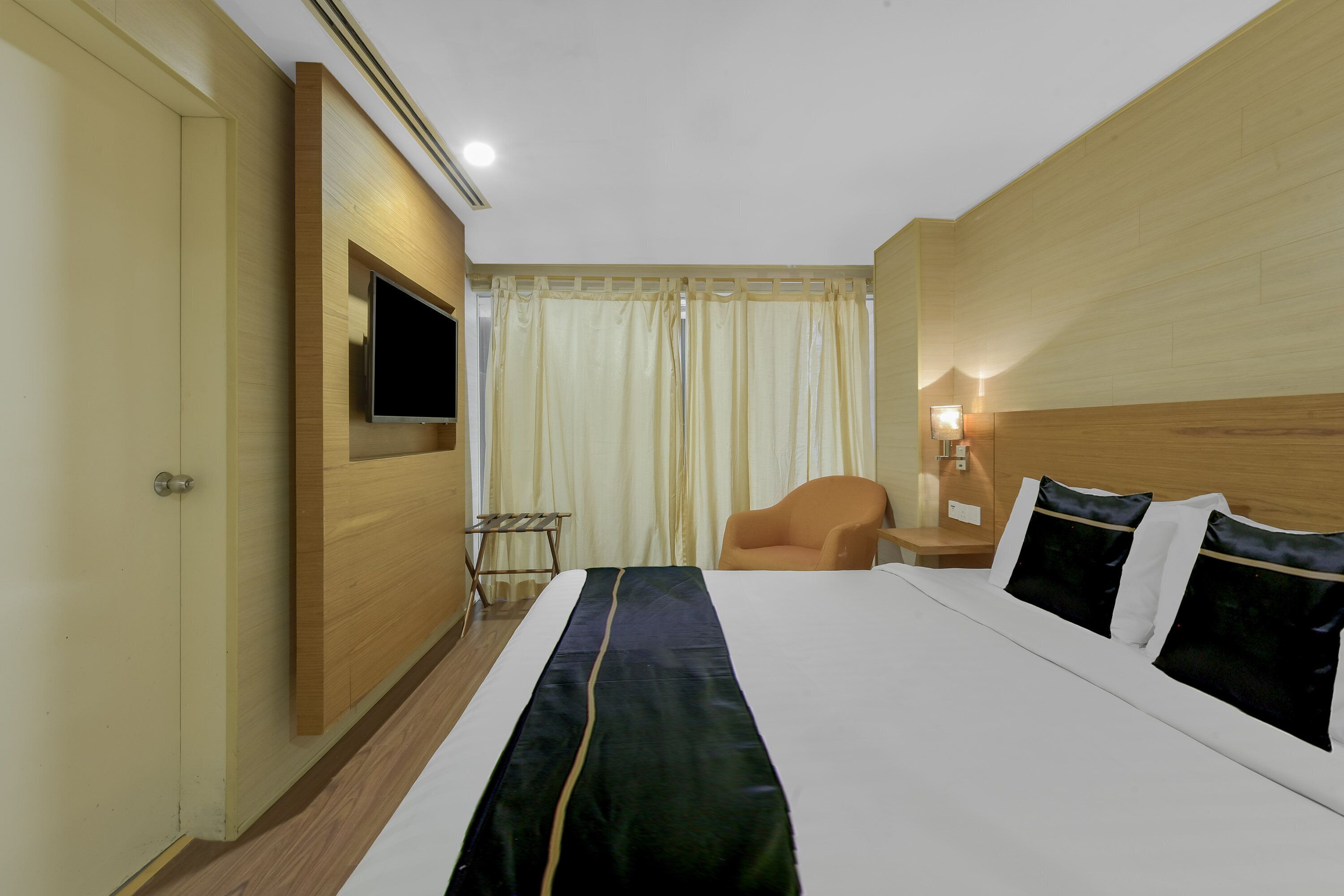Une chambre d’hôtel avec un grand lit, un téléviseur fixé au mur, une chaise, une table de chevet et une fenêtre avec des rideaux.