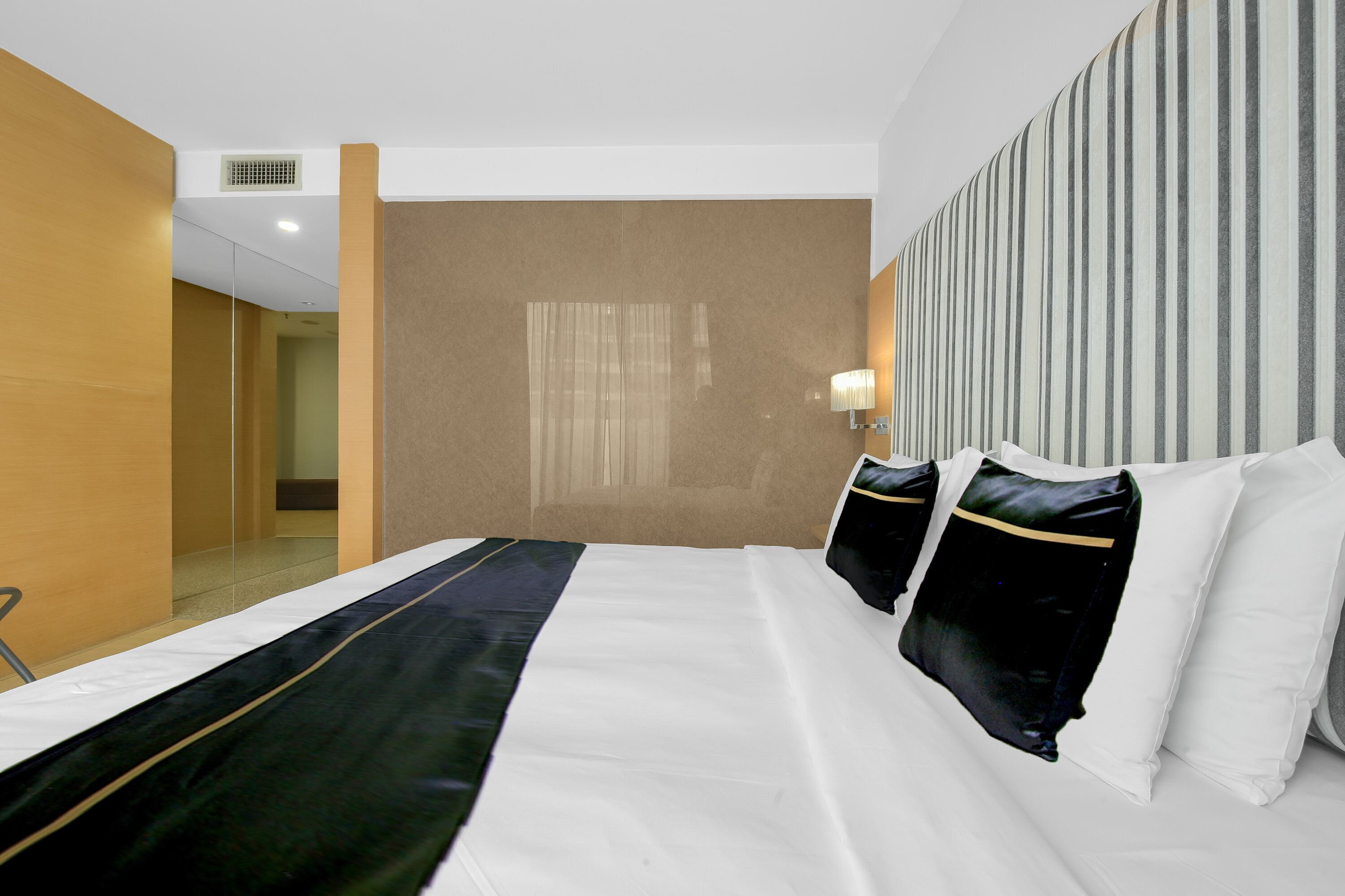 A hotel room with a large bed, a headboard with vertical stripes, a bedside lamp, and a view of the hallway.