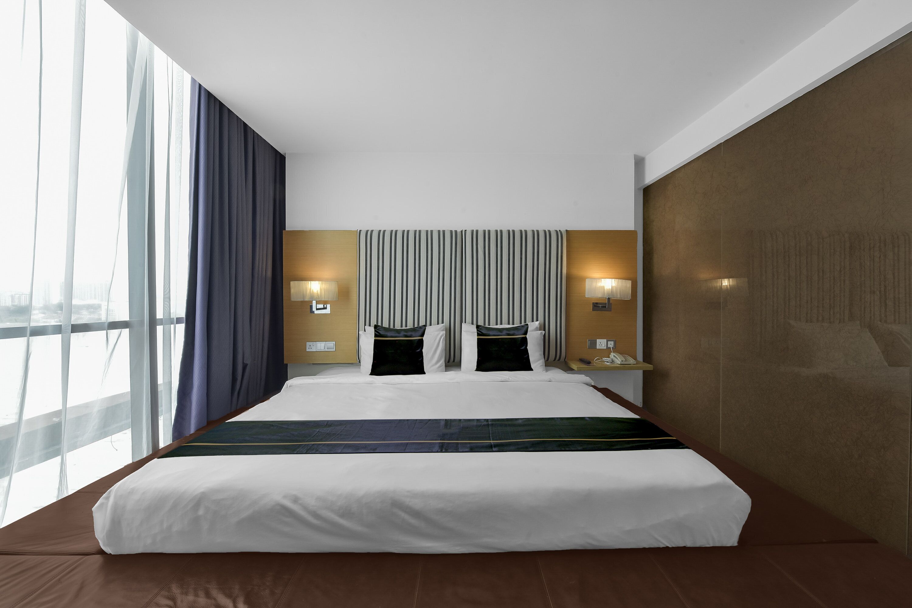 Una habitación de hotel moderna con una cama grande, dos lámparas de noche, vistas de la ciudad a través de amplias ventanas y un cabecero a rayas.