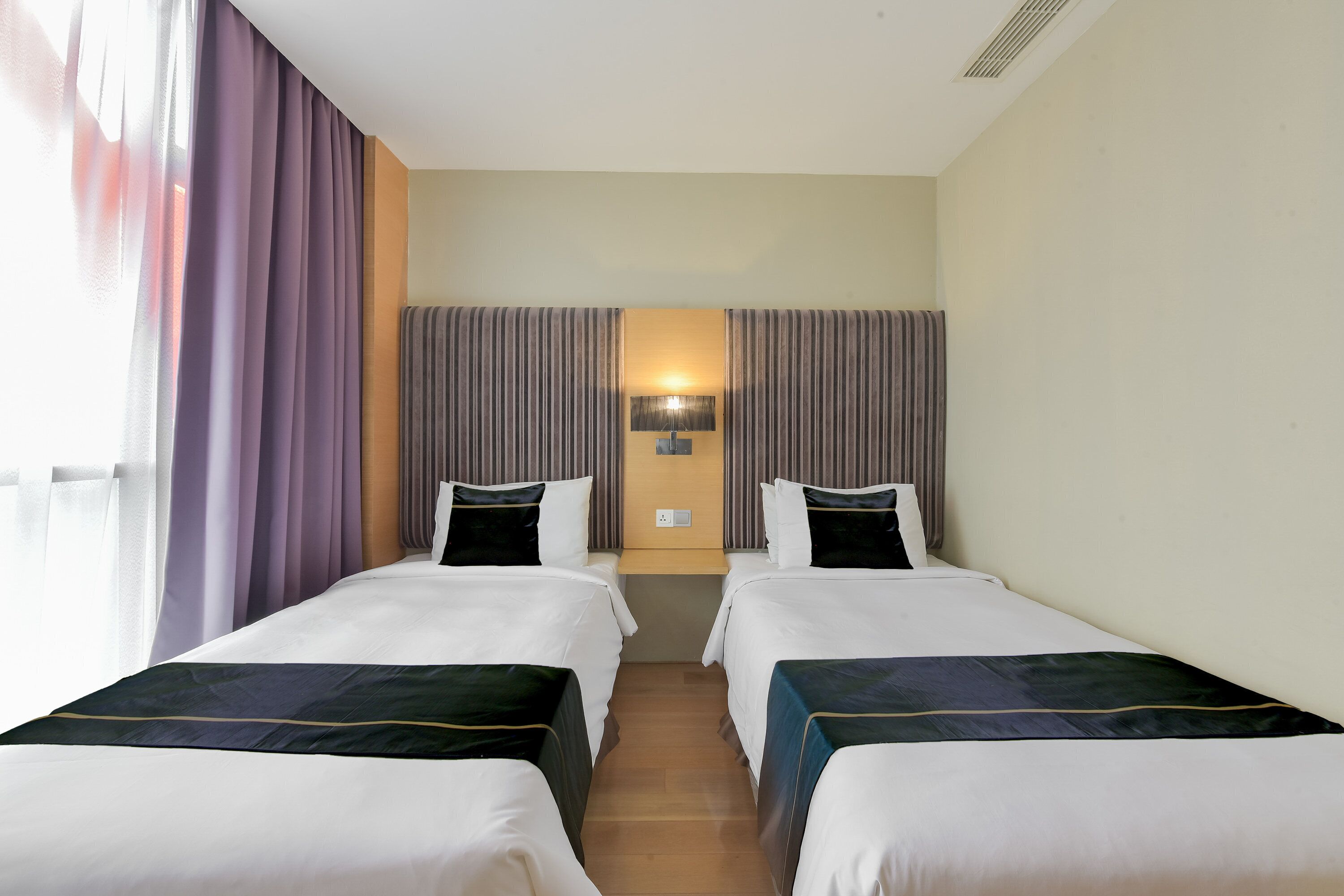 A hotel room with two single beds, a nightstand, a lamp, and a window with curtains.