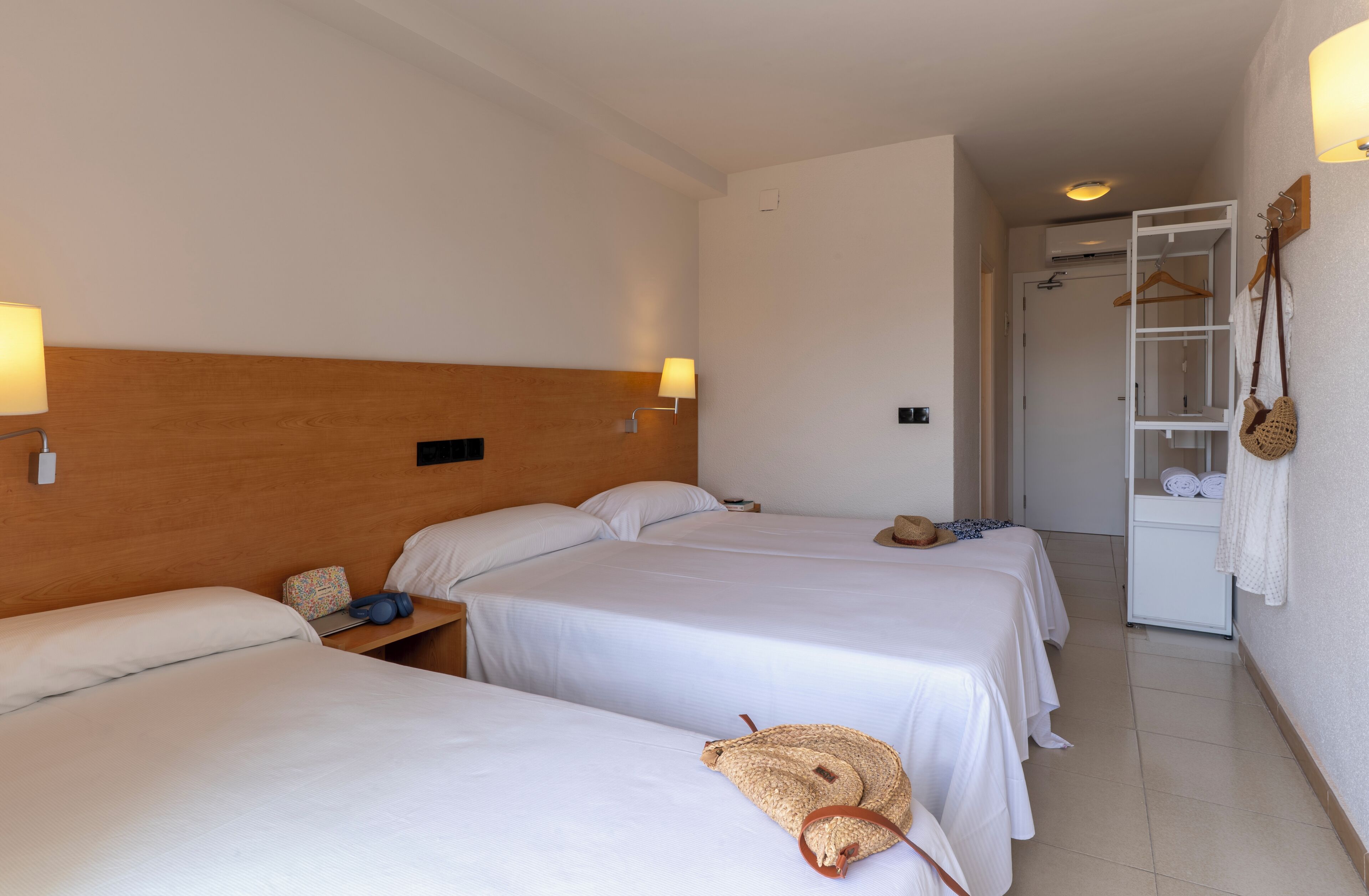 A hotel room with three single beds, a wooden headboard, bedside lamps, a small table with a lamp, and a wardrobe with a hanging basket.