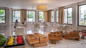 A modern dining area with white chairs and tables, a buffet with various food items, and large windows offering a view of the outdoors. - BAY HOTEL (Roses)