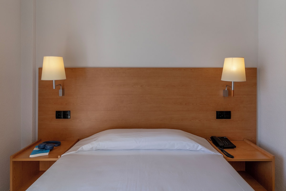 A hotel room with a wooden headboard, two bedside lamps, a bed with white linens, a bedside table with a book, a remote control, and a phone.