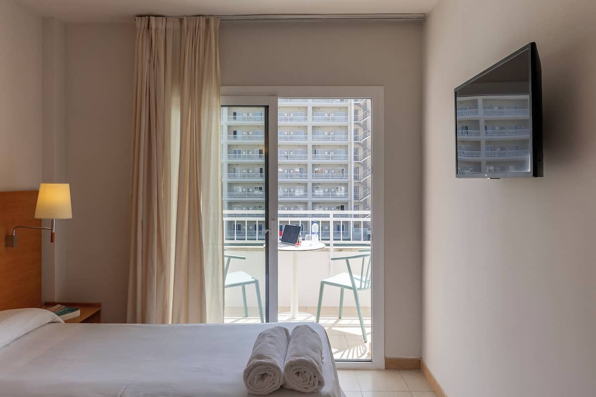 A hotel room with a bed, a nightstand, a lamp, a TV, and a balcony with a table and chairs.