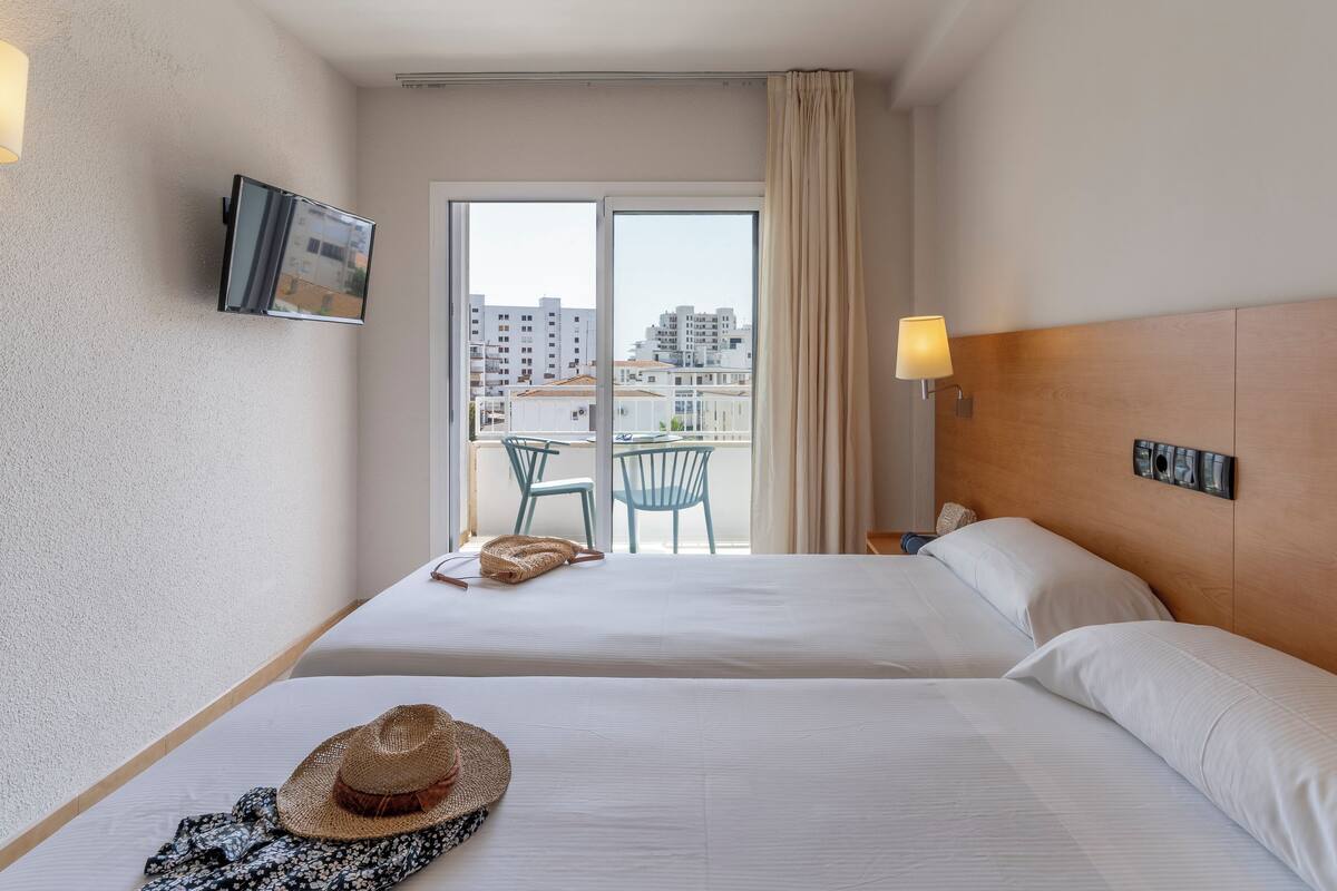A hotel room with two beds, a TV mounted on the wall, a balcony with chairs, and a view of buildings.