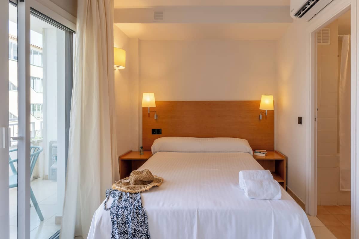 A hotel room with a large bed, two bedside lamps, a wooden headboard, a window with curtains, and a view of the outside.