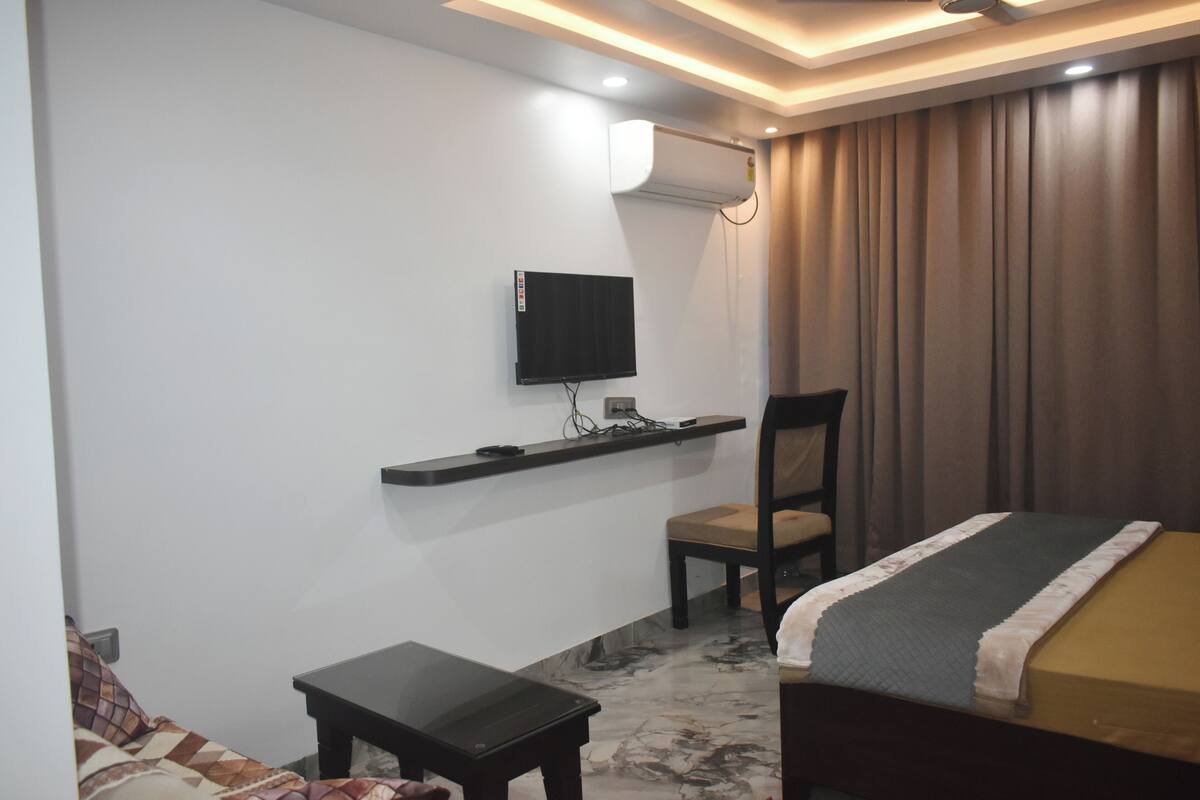 A hotel room with a bed, a small table, a chair, a TV mounted on the wall, and a curtain.
