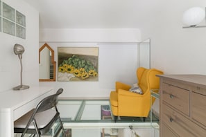 A modern room with a glass partition, a desk, a yellow chair, a wooden dresser, and a painting of sunflowers.