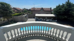 Vista desde un balcón sobre un área de piscina con sillones de descanso y un edificio con techo de tejas al fondo.