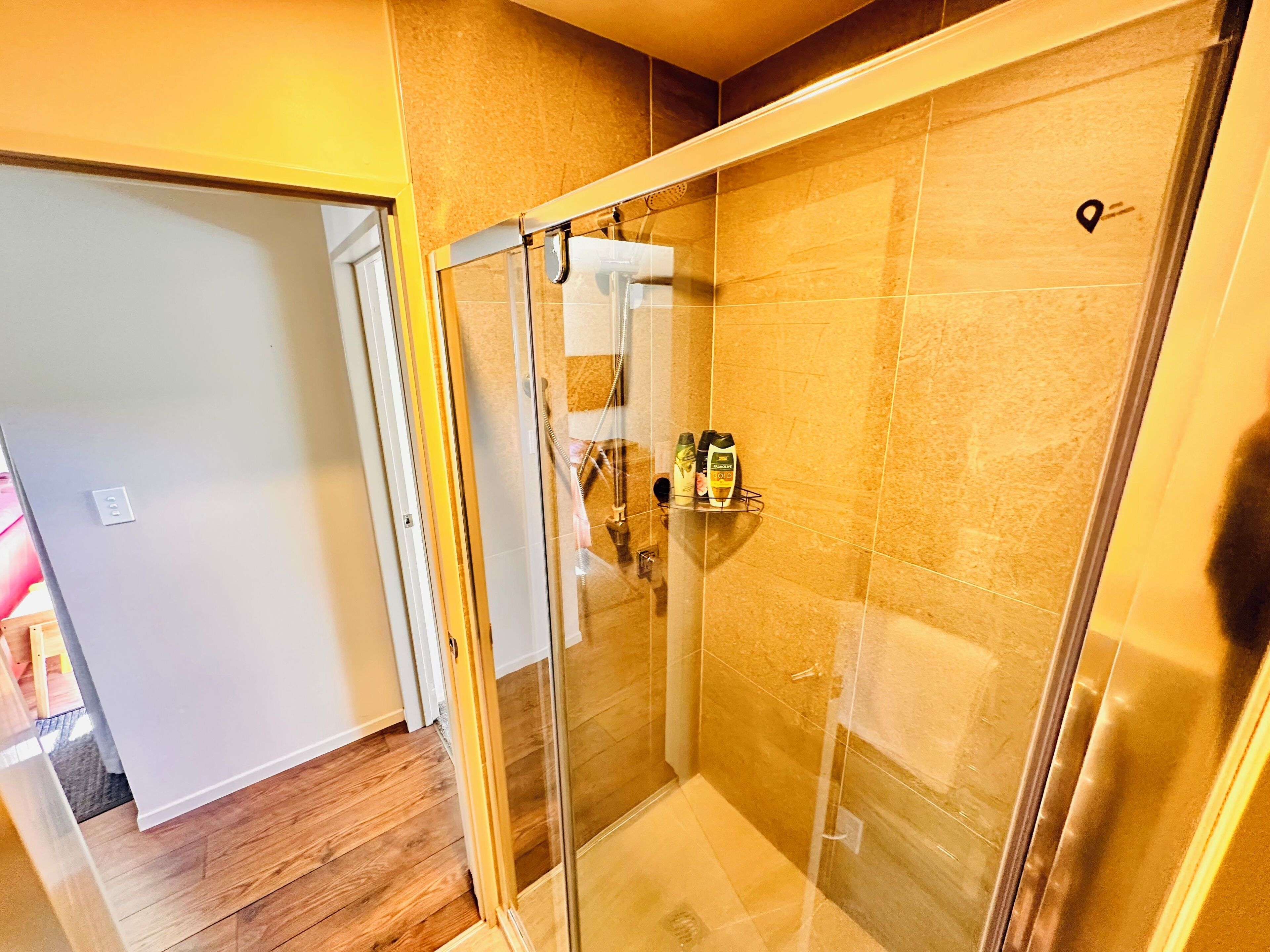 A glass-enclosed shower with a tiled wall, a showerhead, and a soap dispenser on a shelf.