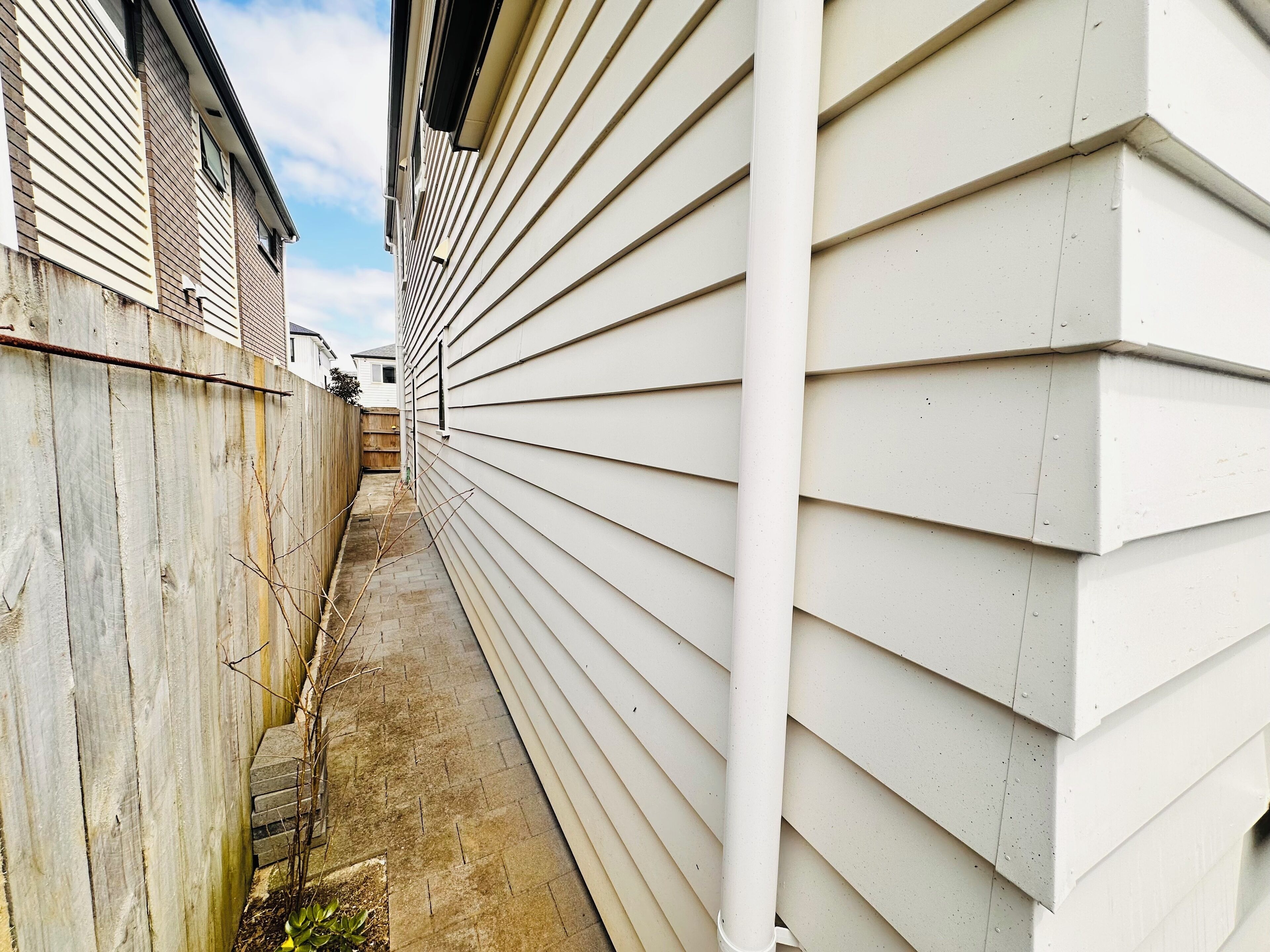 Une ruelle étroite, située entre deux bâtiments recouverts de bardage blanc, avec une clôture en bois d’un côté.