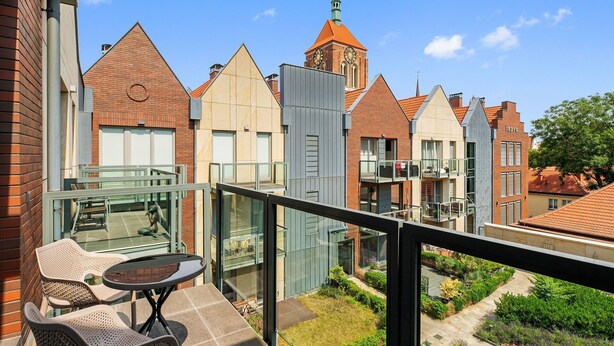A balcony with wicker chairs and a table, overlooking a courtyard with a church tower and adjacent buildings. - Kwartał Kamienic by Downtown Apartments (Gdańsk)