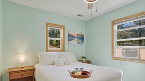 A bedroom with a bed, a nightstand with a lamp, a window with blinds, and a wall-mounted air conditioner.