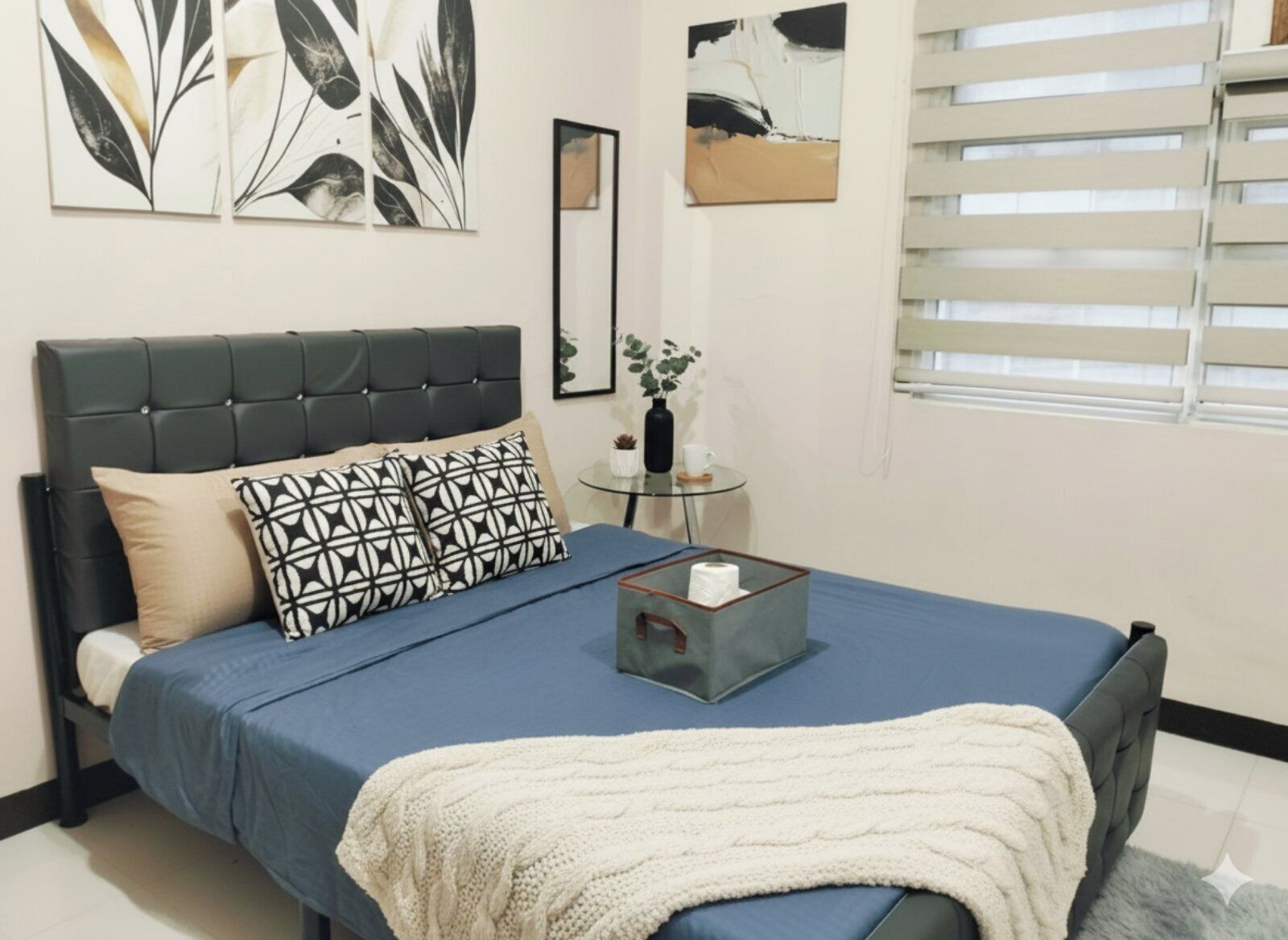 A bedroom with a bed, a bedside table, a window with blinds, and wall art.