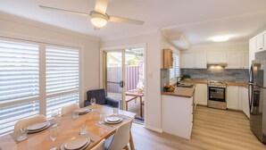 A modern kitchen with white cabinets, a stainless steel refrigerator, and a dining area with a wooden table and chairs. - Crystal by the Bay, 4-6 Crystal Cl - Wifi, Air Con and Coffee machine (Fingal Bay)