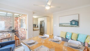 A modern dining area with a brick wall, a kitchen in the background, and a coastal-themed painting on the wall. - Crystal by the Bay, 4-6 Crystal Cl - Wifi, Air Con and Coffee machine (Fingal Bay)