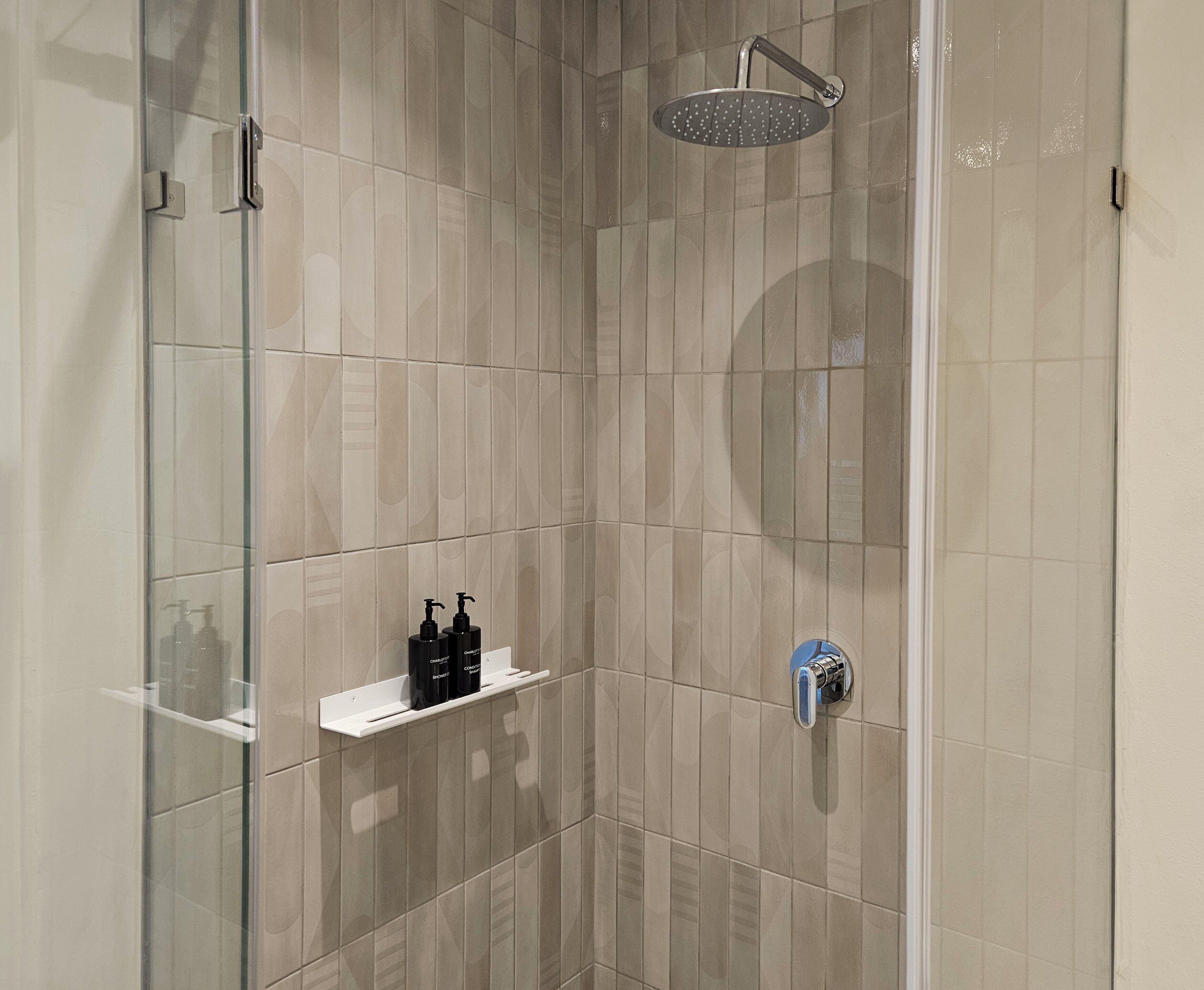 Un espace douche moderne doté d’une porte en verre, de carreaux beiges, d’un pommeau de douche à main et d’une étagère contenant des articles de toilette.